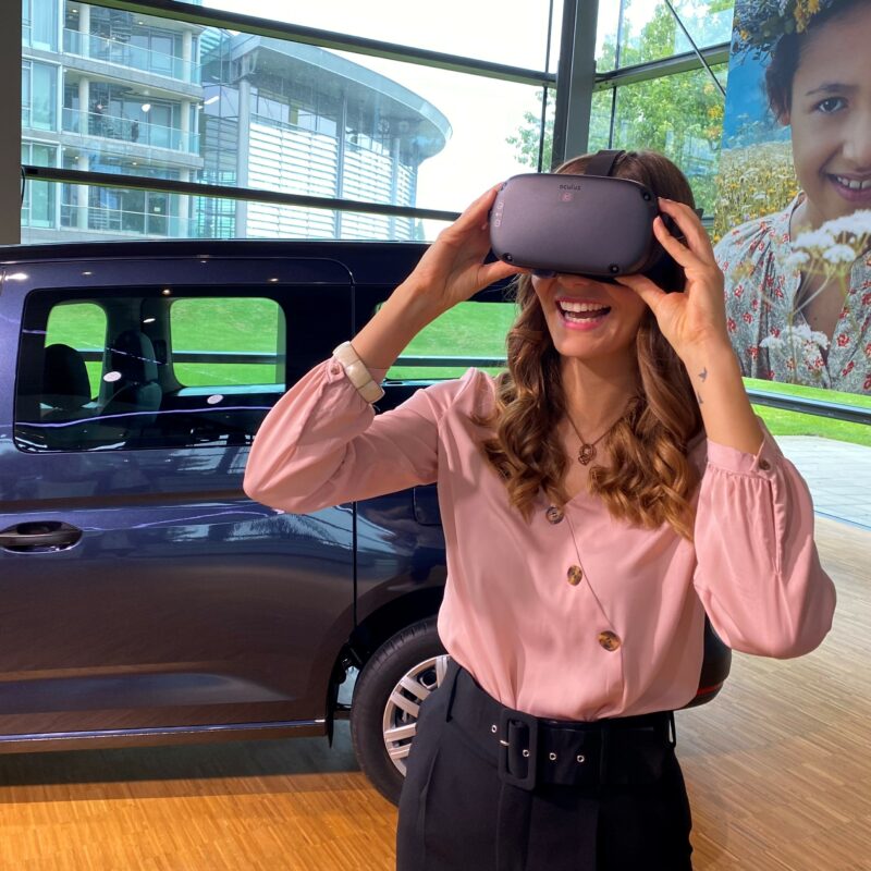 Eine Frau steht vor einem Fahrzeug, trägt eine VR-Brille und lächelt dabei.